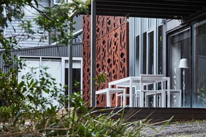 A modern outdoor seating area with a white table and chairs, a red perforated wall, and a glass door leading to a building.