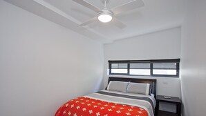 Un dormitorio con cama, mesita de noche, ventana con persianas y ventilador de techo.