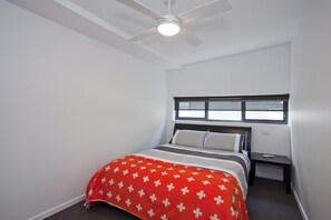 Un dormitori amb un llit, una tauleta de nit, una finestra amb persianes i un ventilador de sostre.