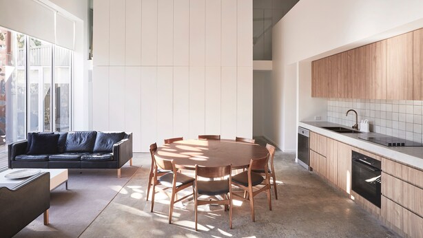 A modern kitchen with wooden cabinets, a dining area with a round table and chairs, and a sofa in a bright room with large windows.