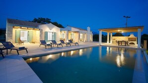A poolside area with lounge chairs, a covered seating area, and a building with open doors and windows. - Exclusive Sea View Villa Madelaine (SPARTIA)