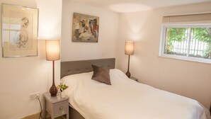A neatly made bed with a pillow, two bedside lamps, a small table with a vase, and a window with blinds.