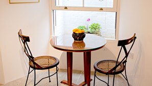 A dining area with a round table and two chairs, a vase with flowers, and a window with a view of greenery.