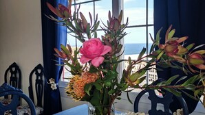 Una mesa de comedor con un florero y protectores de mesa tejidos, frente a una ventana con vista al mar.