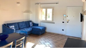 Een kamer met een blauwe modulaire bank, een eettafel met stoelen, een betegelde vloer, een raam met gordijnen en een deur met een beveiligingssysteem.