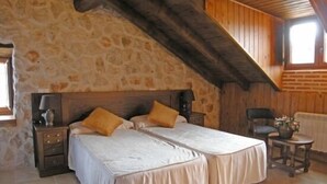 Ein Schlafzimmer mit Steinwand, Holzbalkendecke, einem Doppelbett mit weißen Bettwäsche, zwei Nachttischen mit Lampen und einem kleinen Tisch mit einer Pflanze.