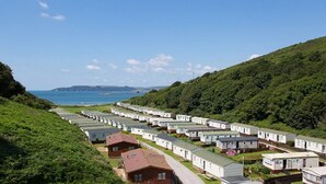 A caravan park with rows of caravans, a few houses, and a view of the sea and hills in the background. - 2 Bed Caravan, South Devon Coast nr Plymouth, 200m to Beach + Indoor Heated Pool (PLYMOUTH)