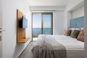 Une chambre d’hôtel moderne avec un grand lit, un téléviseur fixé sur une étagère en bois et un balcon offrant une vue sur la mer.