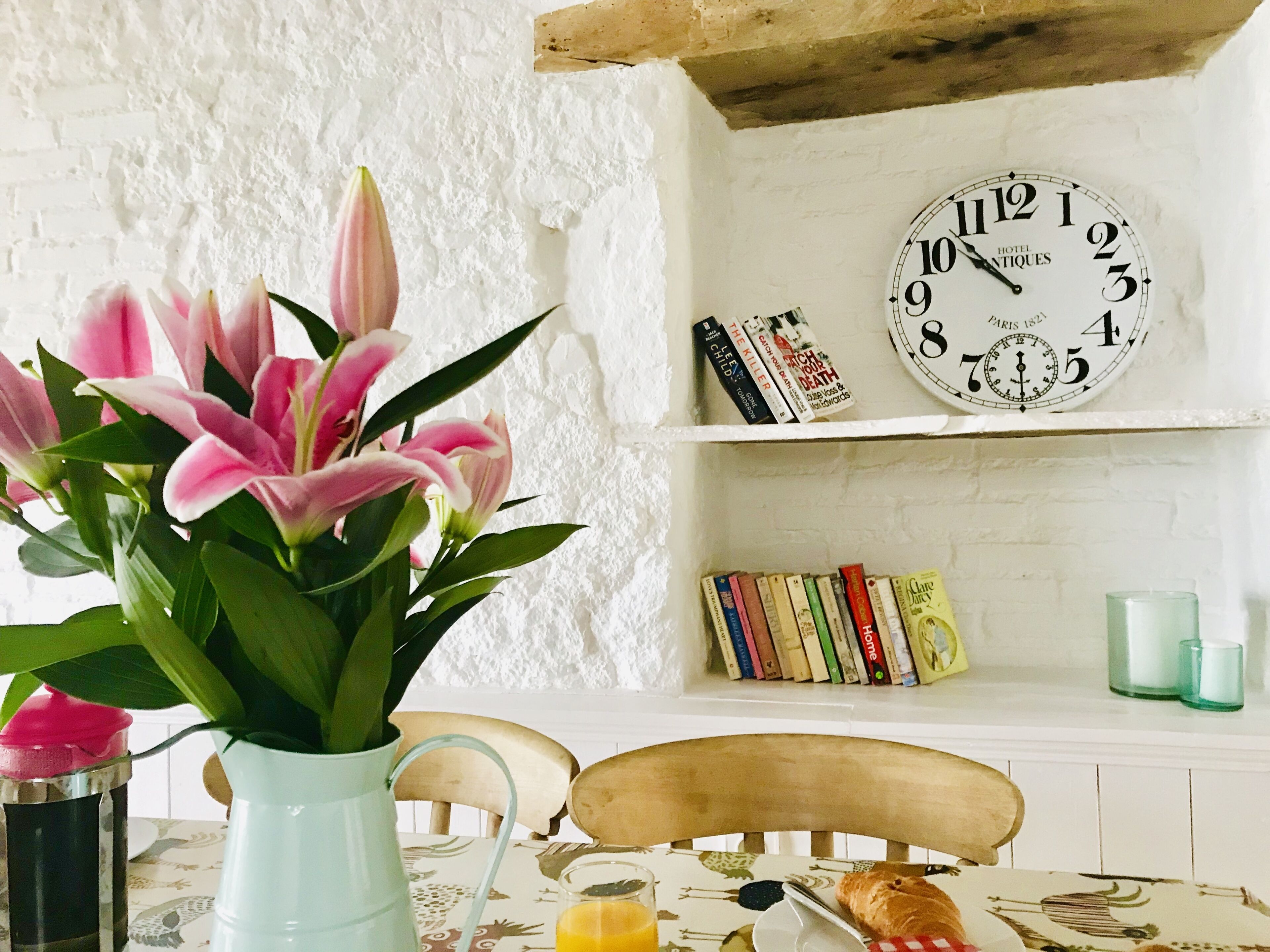 Una mesa con un jarrón de lirios rosados, un vaso de jugo de naranja y un croissant. Una silla de madera está colocada detrás de la mesa. Se ve un muro de ladrillo blanco con un reloj blanco y estanterías en el fondo.