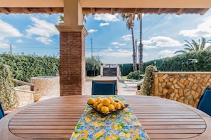 An outdoor patio with a table set for a meal, a stone wall, and a view of a gate and greenery.