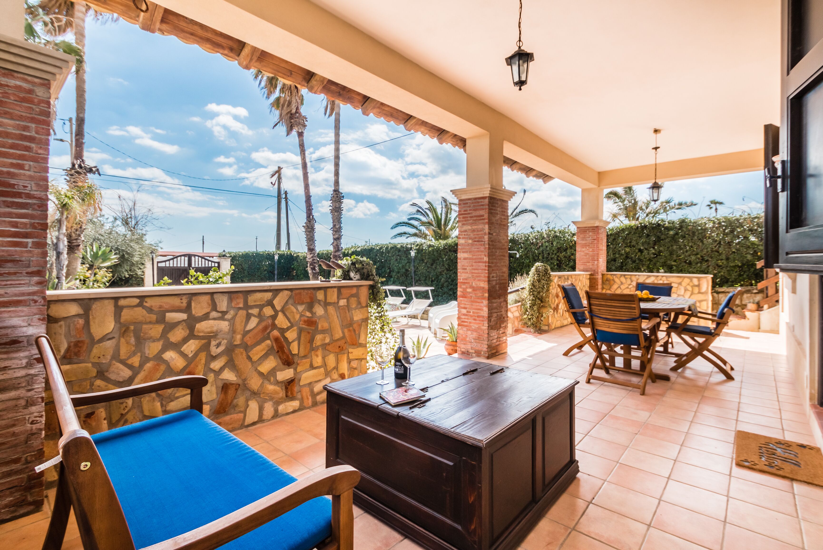 Un patio coperto con un muro in pietra, un tavolo di legno con sedie e una poltrona imbottita blu.