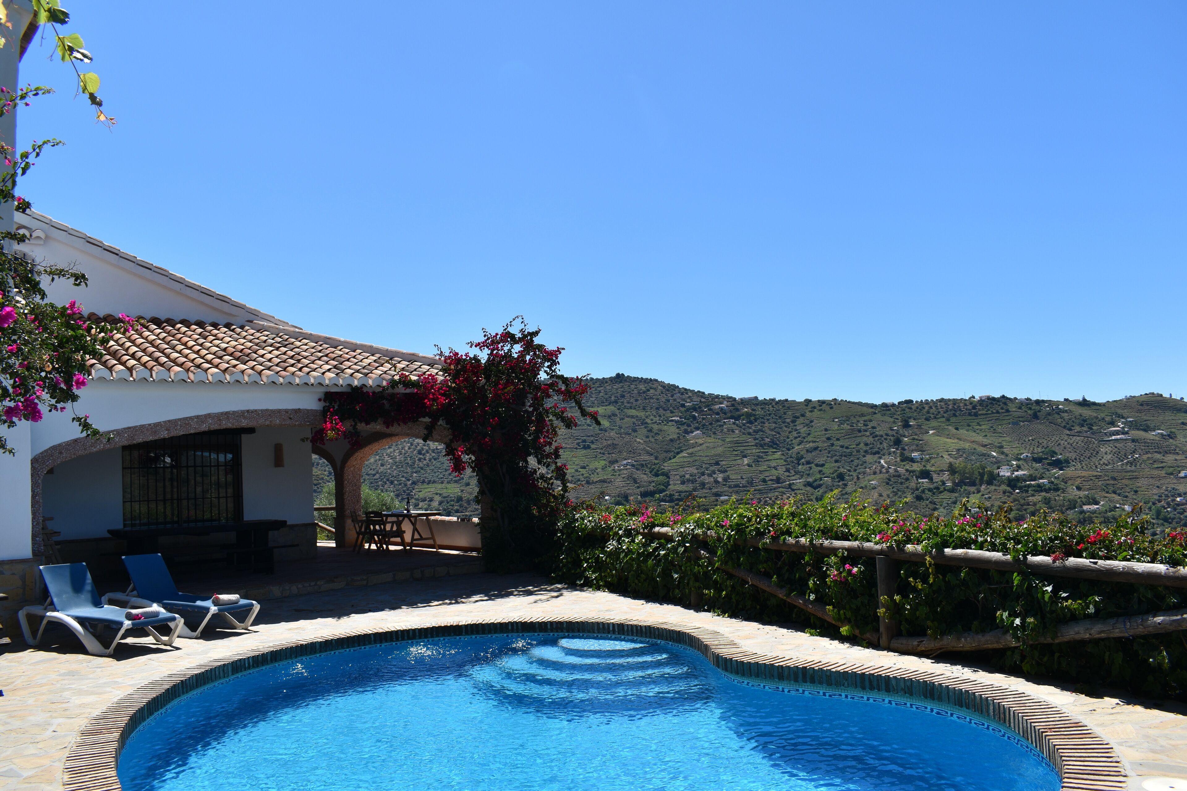 Une piscine avec des chaises longues, une pergola ornée de fleurs et une vue sur des collines verdoyantes.