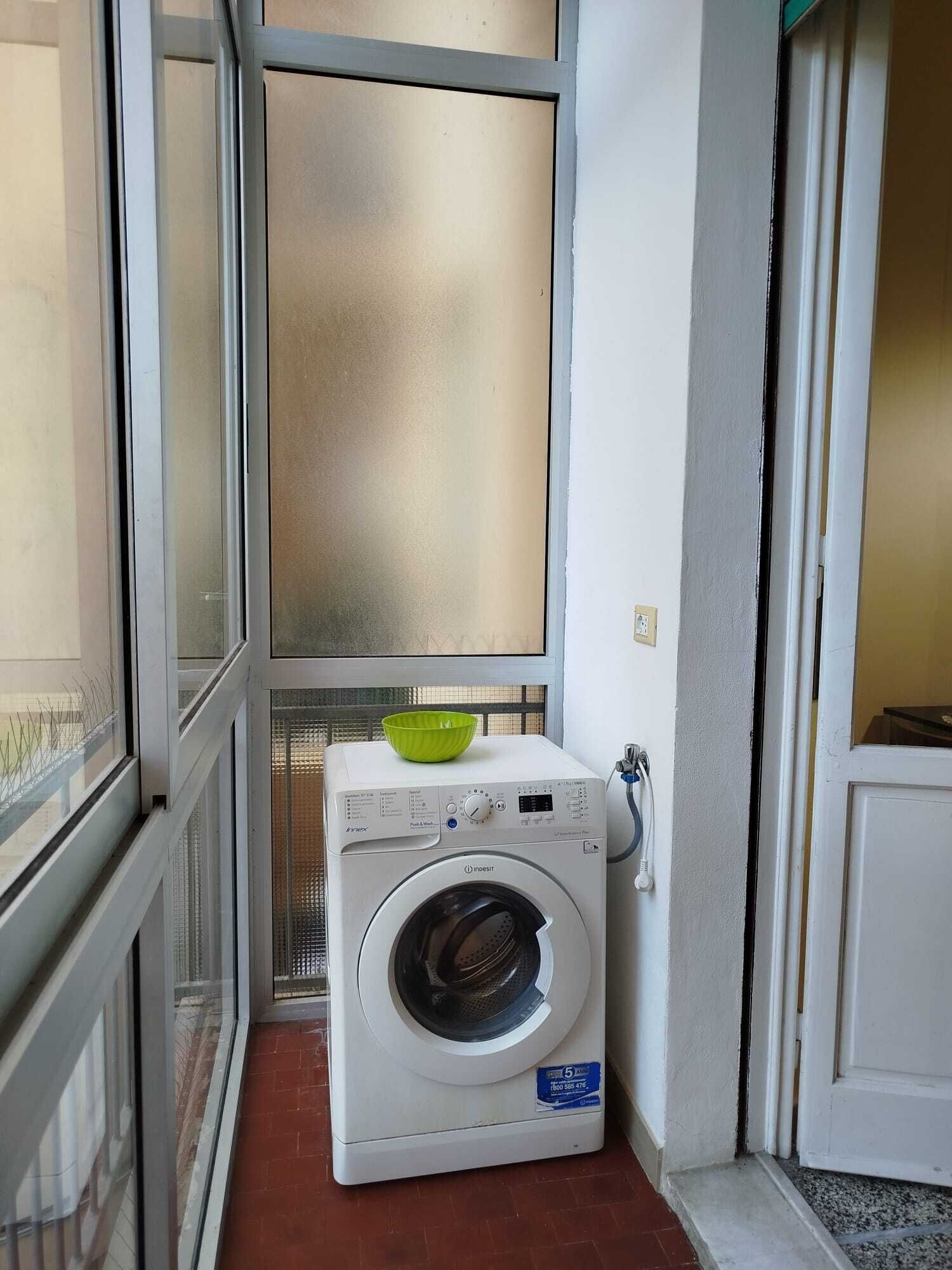 Une petite buanderie équipée d’un lave-linge à chargement frontal, avec un saladier vert posé sur le dessus, et une fenêtre à vitre dépolie.