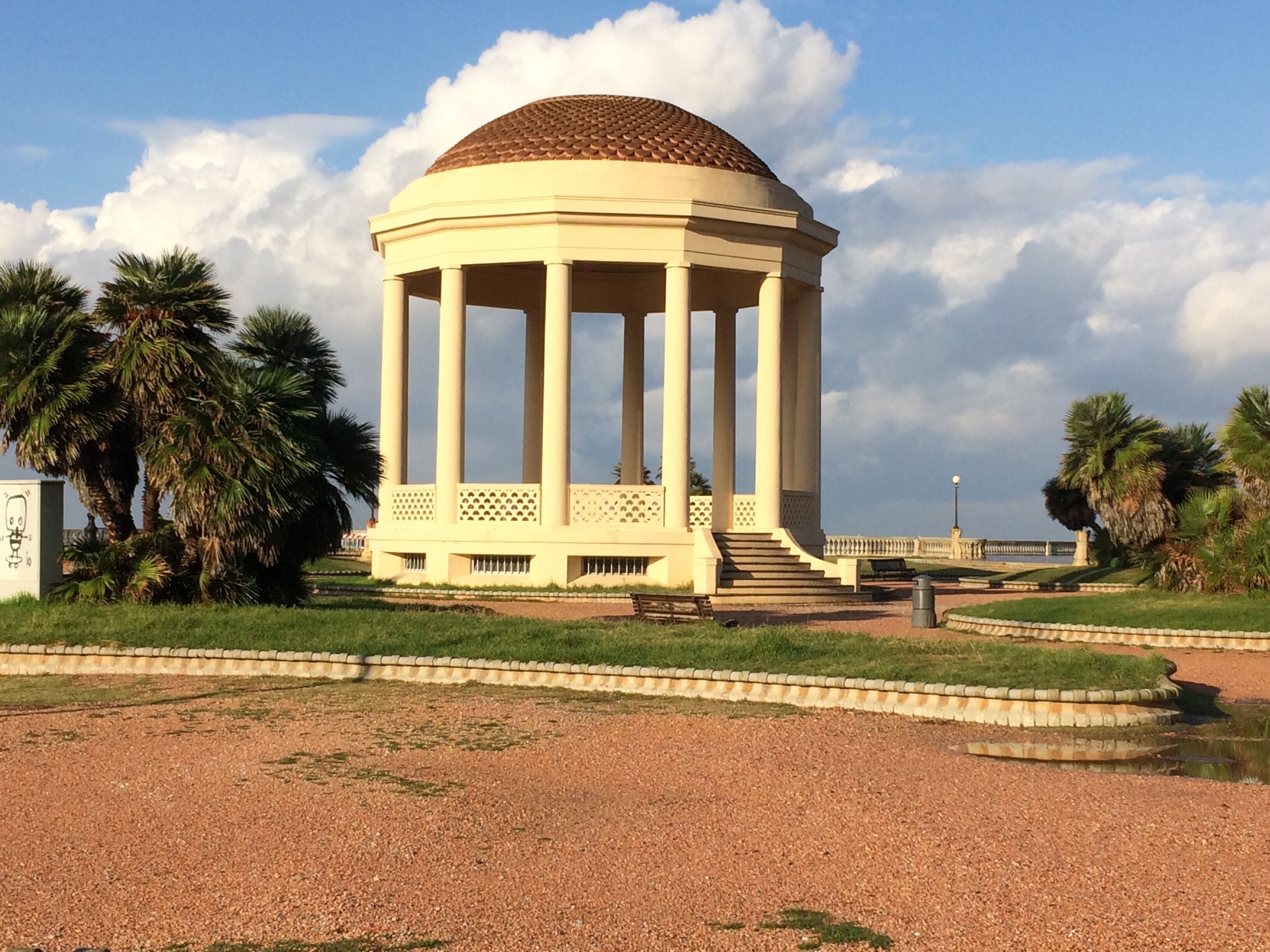 A pavilion with a dome roof, surrounded by palm trees and a gravel path.