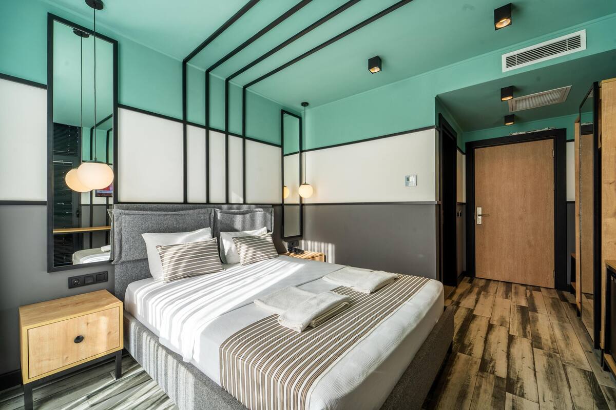 A modern hotel room with a large bed, a bedside table, a wardrobe, and a mirror.