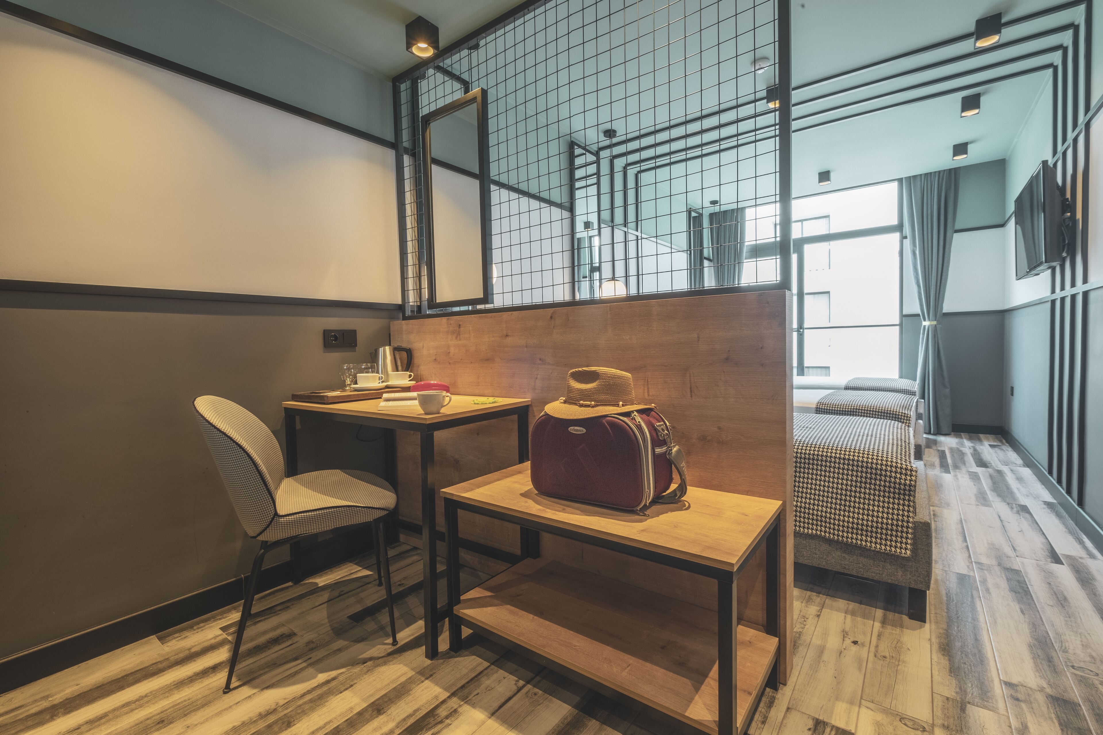 Habitación de hotel moderna con suelo de madera, una cama individual, una mesita con una silla, una maleta y un ventanal grande con cortinas.