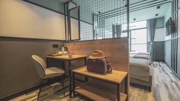 Habitación de hotel moderna con suelo de madera, una cama individual, una mesita con una silla, una maleta y un ventanal grande con cortinas.