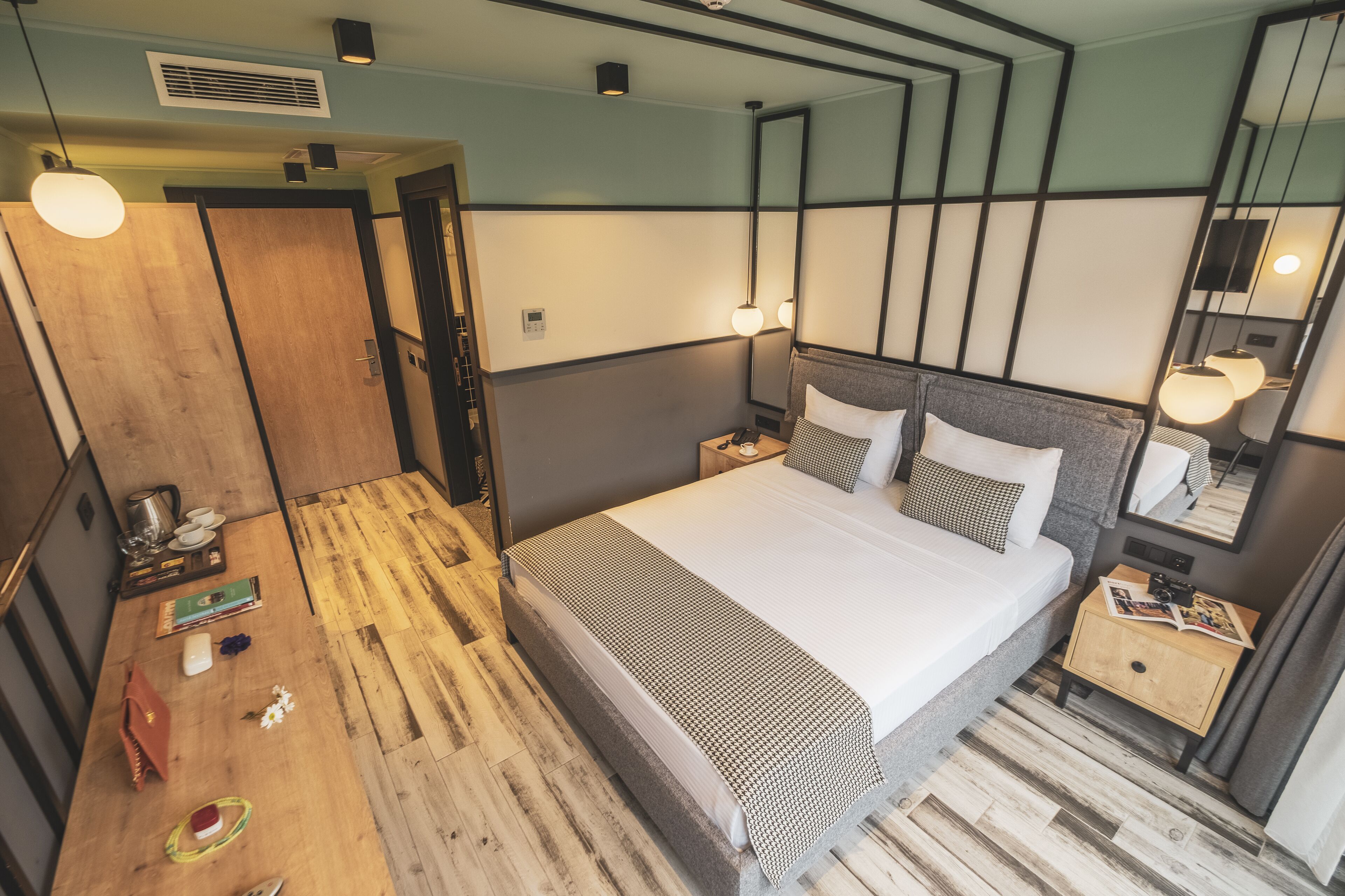 A modern hotel room with a wooden floor, a neatly made bed, a bedside table with a lamp, and a small wooden nightstand.