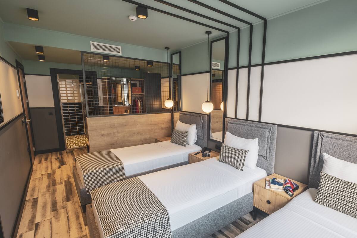 A hotel room with two single beds, a wooden floor, a mirror, and a small bedside table with a book and a flag.