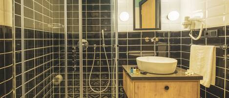 Baño con paredes de azulejos blancos y negros, cabina de ducha de vidrio, tocador de madera con lavabo y espejo con iluminación.