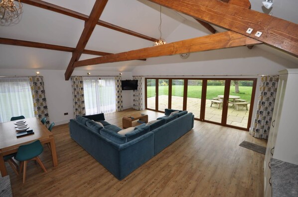 A spacious living room with a large sofa, wooden dining table, and a view of a garden through glass doors.