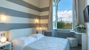 A hotel room with a bed, bedside tables, a window with a view, a radiator, and a TV on the wall. - Villa Leonardo 30+6, Emma Villas (Figline e Incisa Valdarno)