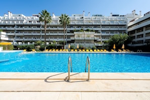 A swimming pool with a clear blue water, surrounded by yellow lounge chairs and umbrellas, with a multi-story building in the background.
