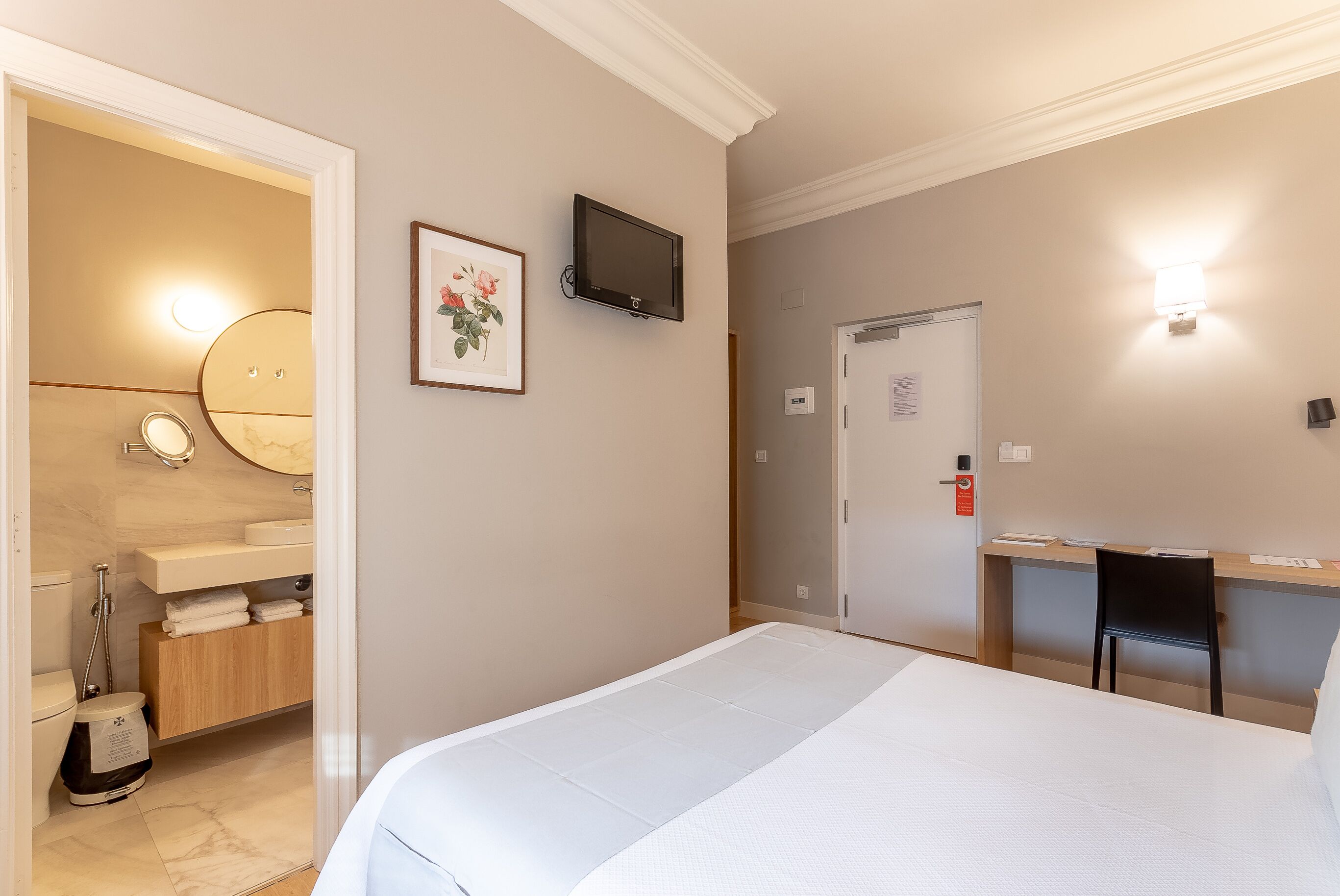Habitación de hotel con cama, escritorio y silla, televisión, baño con espejo y un cuadro en la pared.