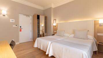 A hotel room with a double bed, a wooden desk, a wardrobe, and a bedside table with a lamp.