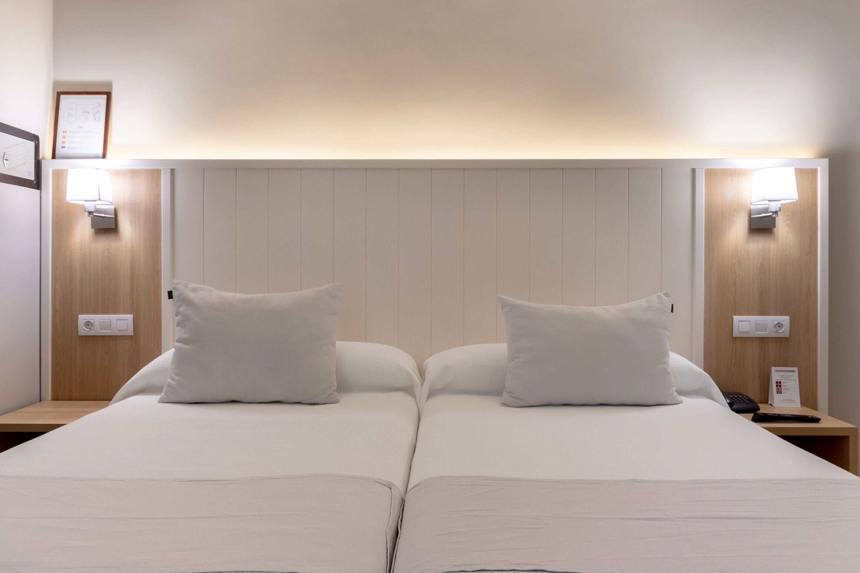 Una cama doble con sábanas blancas y dos almohadas, una mesita de noche con lámpara, y una lámpara de pared.