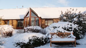 A house with a snow-covered garden, a wooden bench, and a stone facade.