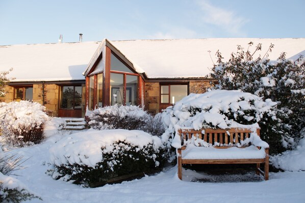 Une maison avec un jardin enneigé, un banc en bois et une façade en pierre.