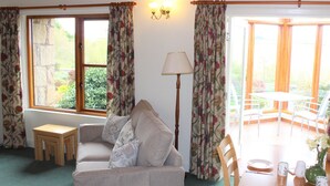 A cozy living room with a sofa, a small table, and a dining table with chairs. There are large windows with curtains, a lamp, and a view of the outdoors.