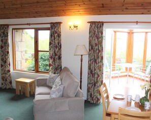 Un salon chaleureux avec un canapé, une petite table et une table à manger avec des chaises. De grandes fenêtres sont agrémentées de rideaux, une lampe est présente, et la vue donne sur l’extérieur.
