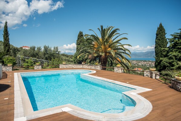 A swimming pool with a wooden deck, surrounded by trees and a view of a coastal town. - Villa Panorama with Sea View and common Swimming Pool (Selianitika)