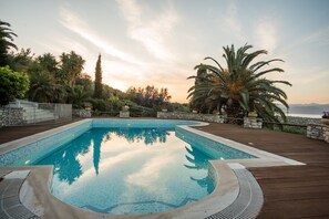 A rectangular swimming pool with a wooden deck surrounding it, set against a backdrop of trees and a clear sky. - Villa Panorama with Sea View and common Swimming Pool (Selianitika)