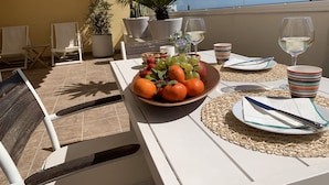 Une table dressée pour un repas, avec un saladier de fruits, des verres à vin et une tasse à rayures, sur une terrasse agrémentée de chaises et de plantes en pot.