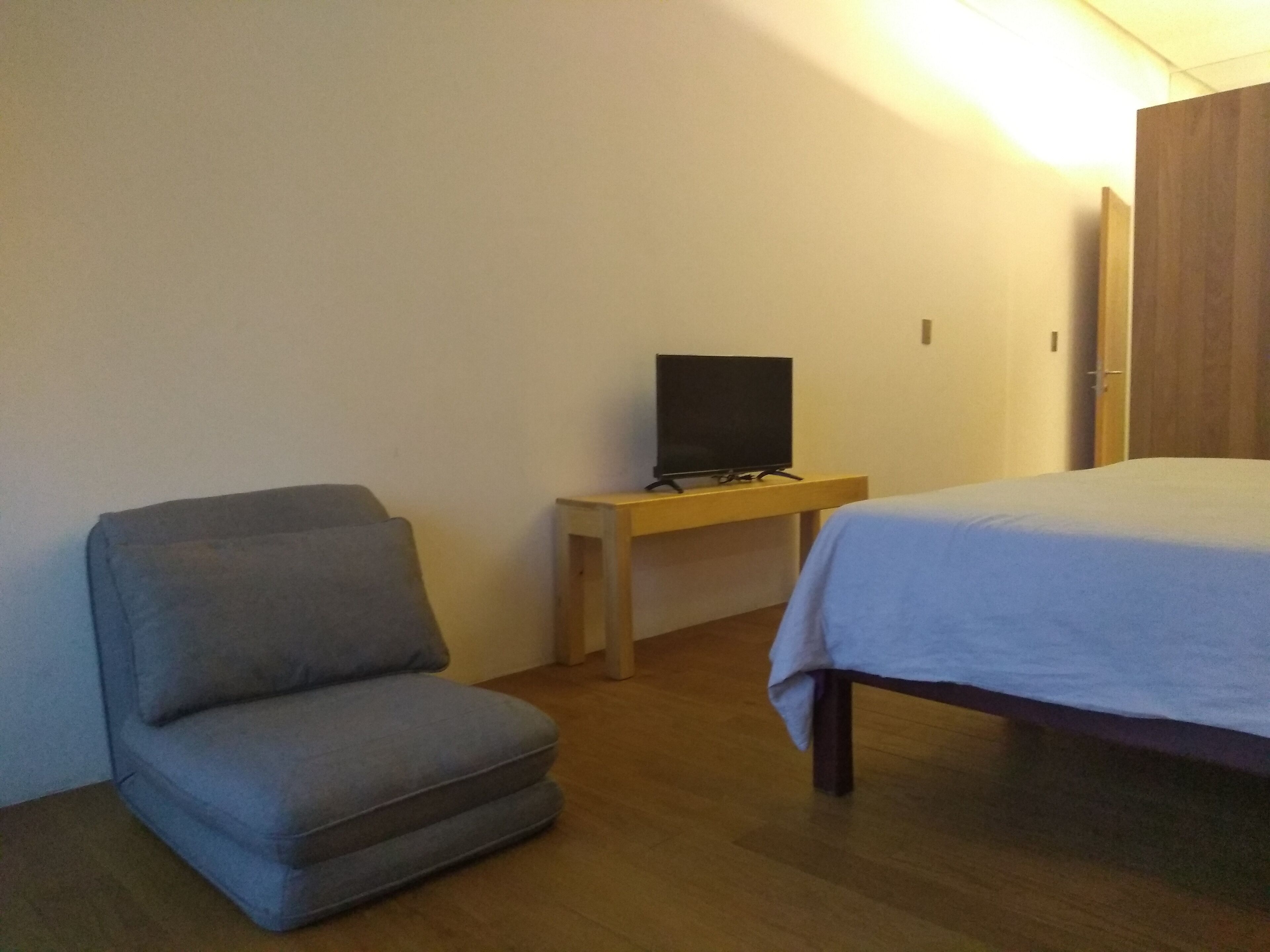 A small, simple room with a bed, a chair, and a TV on a wooden stand.