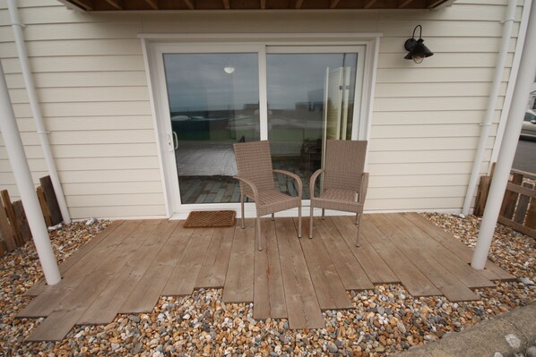 A covered patio with two wicker chairs, a wooden deck, and a glass door leading to a backyard. - Beautiful Seafront Beach House (Selsey)