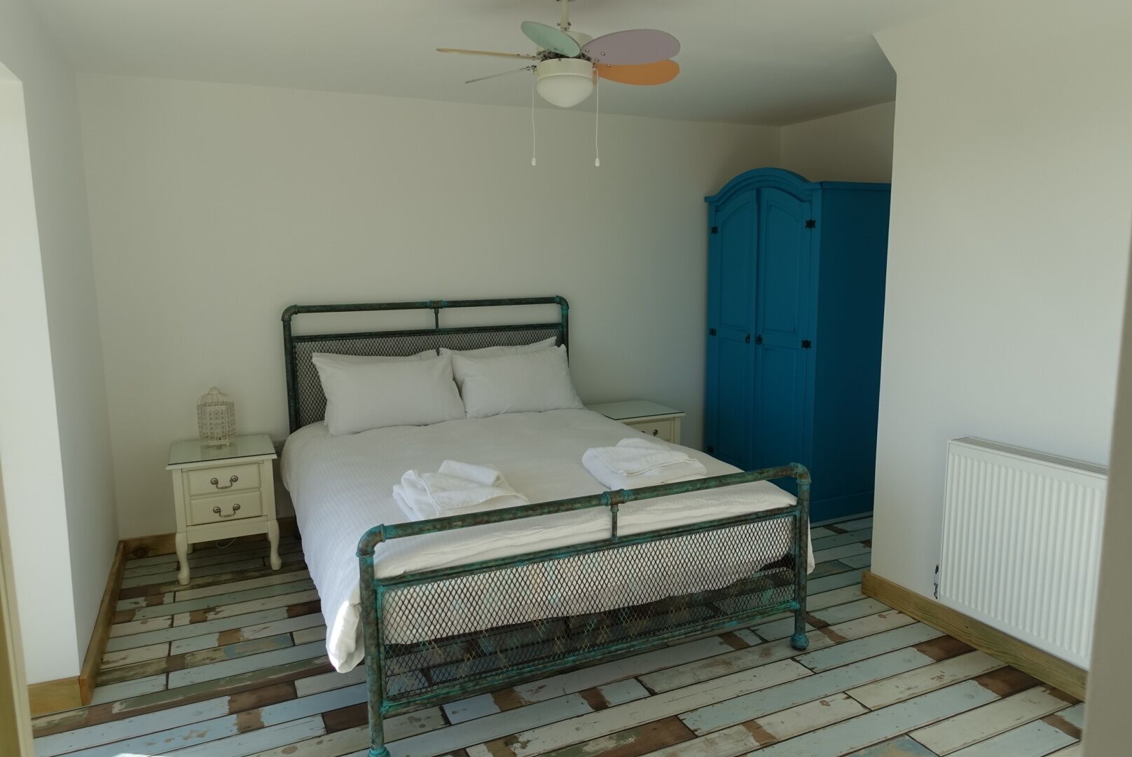 A bedroom with a bed, a nightstand, a blue wardrobe, a ceiling fan, and a radiator.