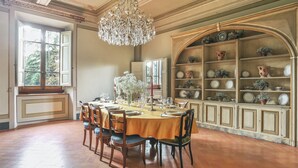 A dining room with a large table set for six, surrounded by chairs. A chandelier hangs from the ceiling, and a bookshelf with decorative plates and vases is to the right. - Villa Franchetti - Luxurious Florentine Villa with Swimming Pool (Firenze)