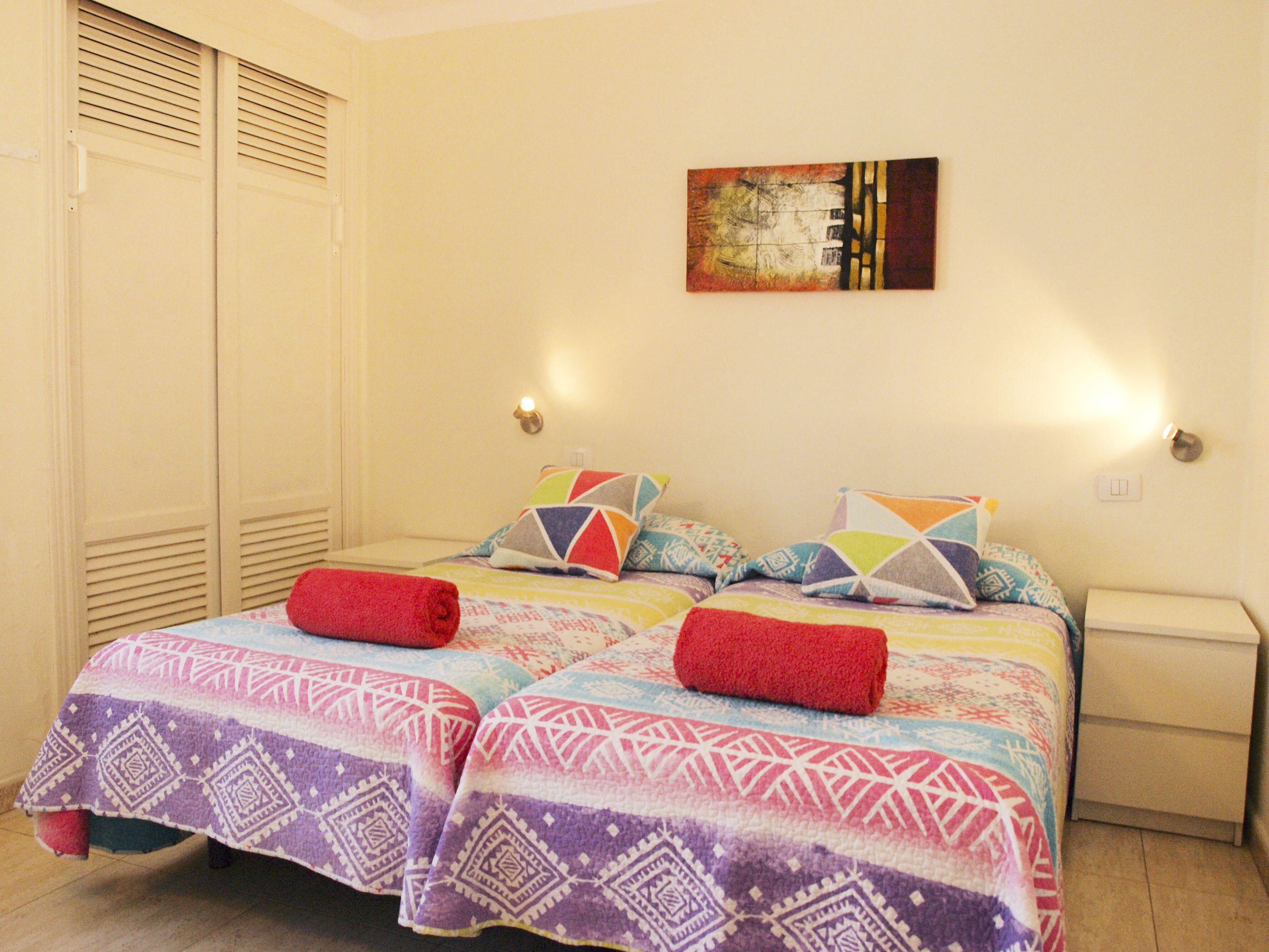 A bedroom with two beds, colorful bedding, a painting on the wall, and a nightstand with a lamp.