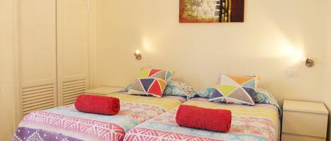 A bedroom with two beds, colorful bedding, a painting on the wall, and a nightstand with a lamp.