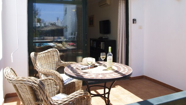 Une terrasse ensoleillée, meublée de meubles en osier, avec une table sur laquelle se trouvent une bouteille et des verres, et une porte-fenêtre coulissante donnant sur un espace intérieur.