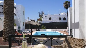 Un espace aménagé au bord de la piscine, avec vue sur un bâtiment, une table avec une bouteille et des verres, et une chaise en osier.