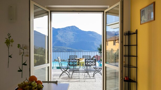 A room with a view of mountains and a pool. There is a table with a bowl of fruit and a chair.