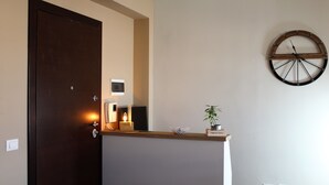Una habitación con una puerta de madera, un reloj en la pared, un pequeño estante con una planta y una lámpara encendida.