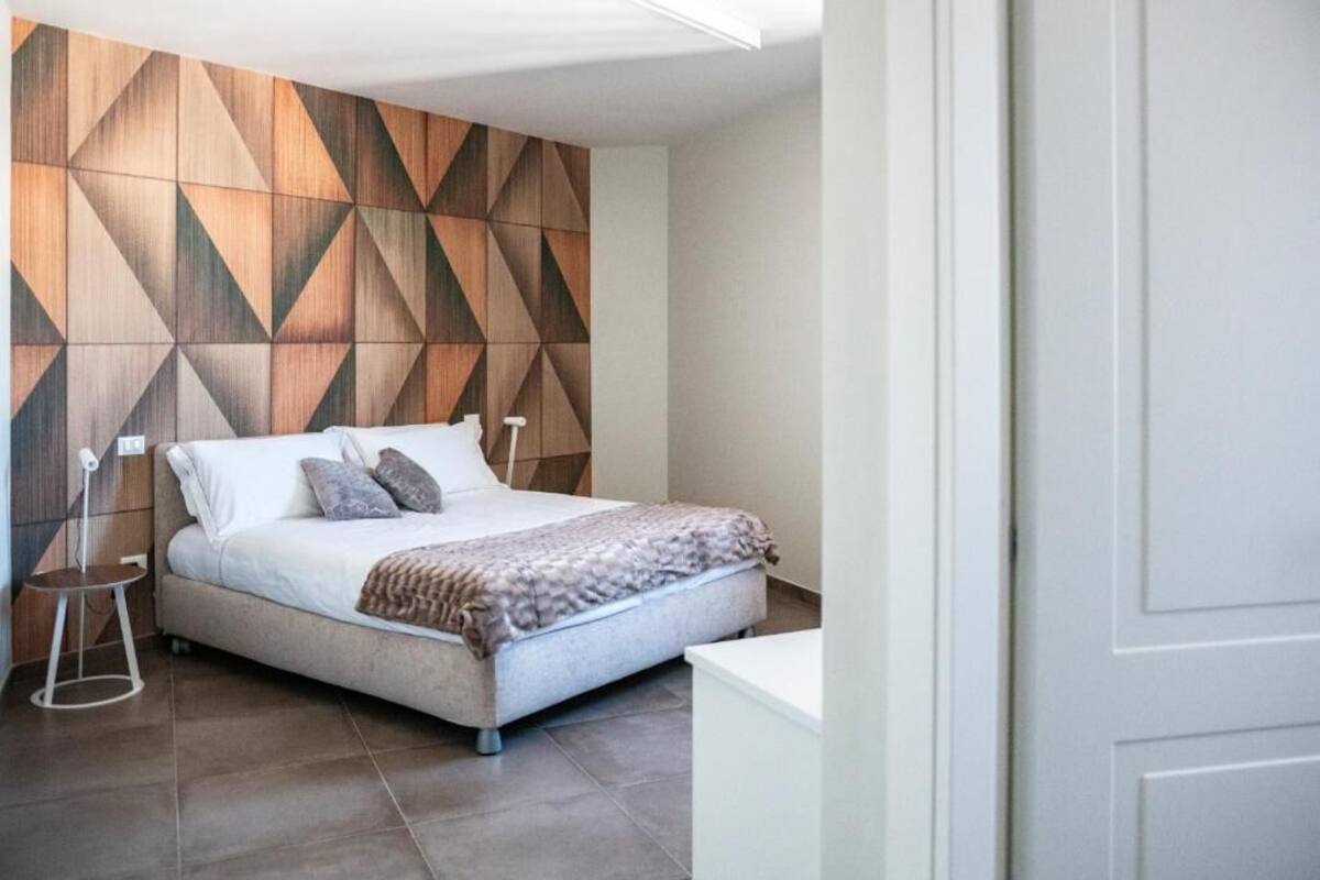 A modern bedroom with a geometric-patterned wall, a bed with pillows, a bedside table, and a nightstand.