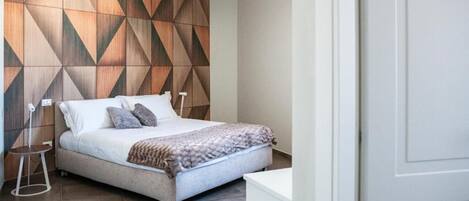 A modern bedroom with a geometric-patterned wall, a bed with pillows, a bedside table, and a nightstand.