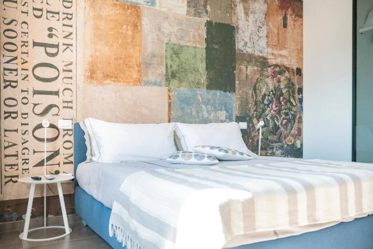 A bed with white bedding and striped pillows, a small white side table, and a colorful abstract wall art piece.